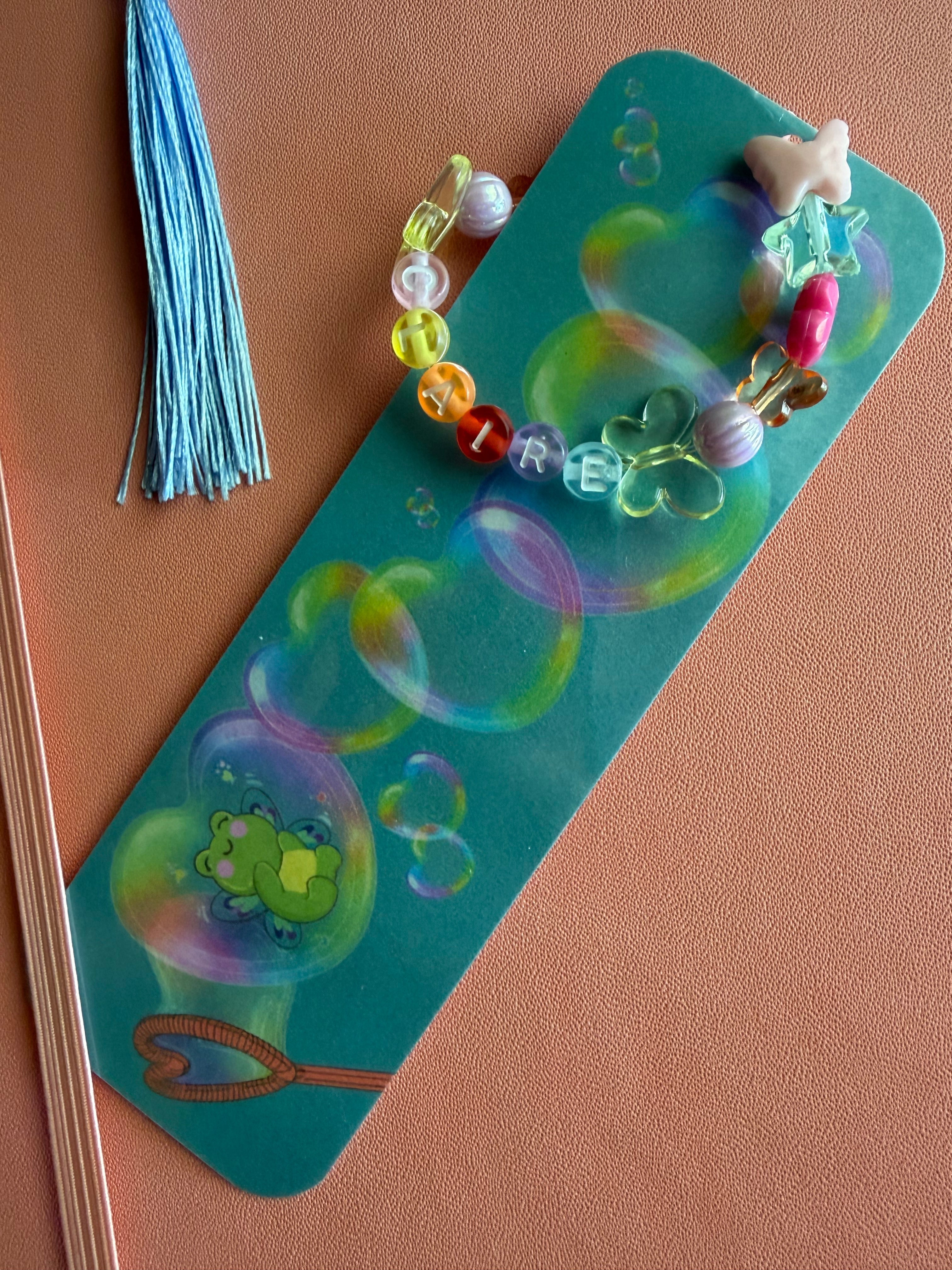 A decorative bookmark with a blue-green background, printed with colorful bubble illustrations and a small frog motif, adorned with multicolored beads and a light blue tassel, displayed on a dusty pink journal.