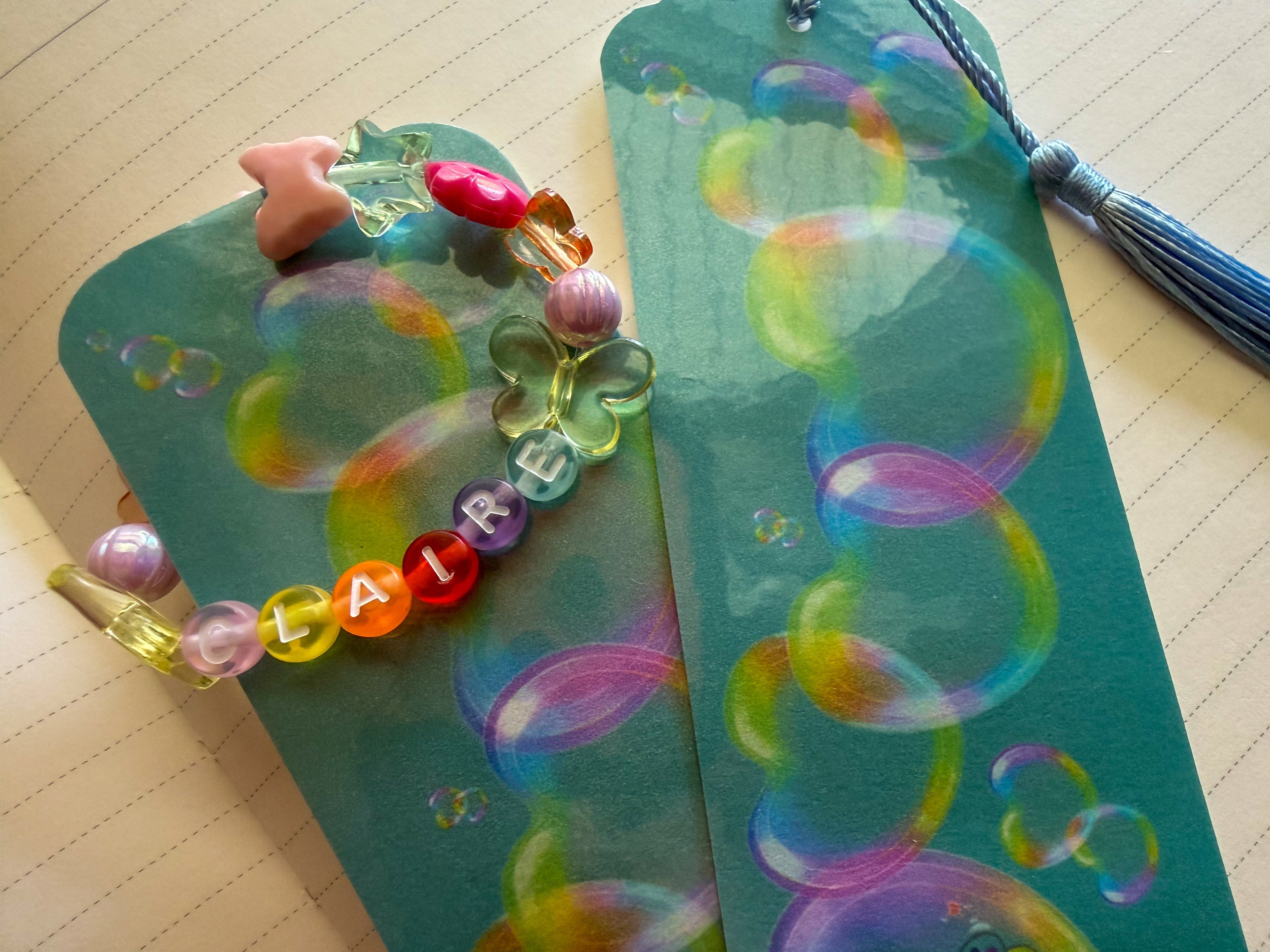 A close up photograph of colorful beaded charm, displayed on a teal bookmark decorated with bubble illustrations, next to a matching bookmark with a blue tassel on a lined notebook page.