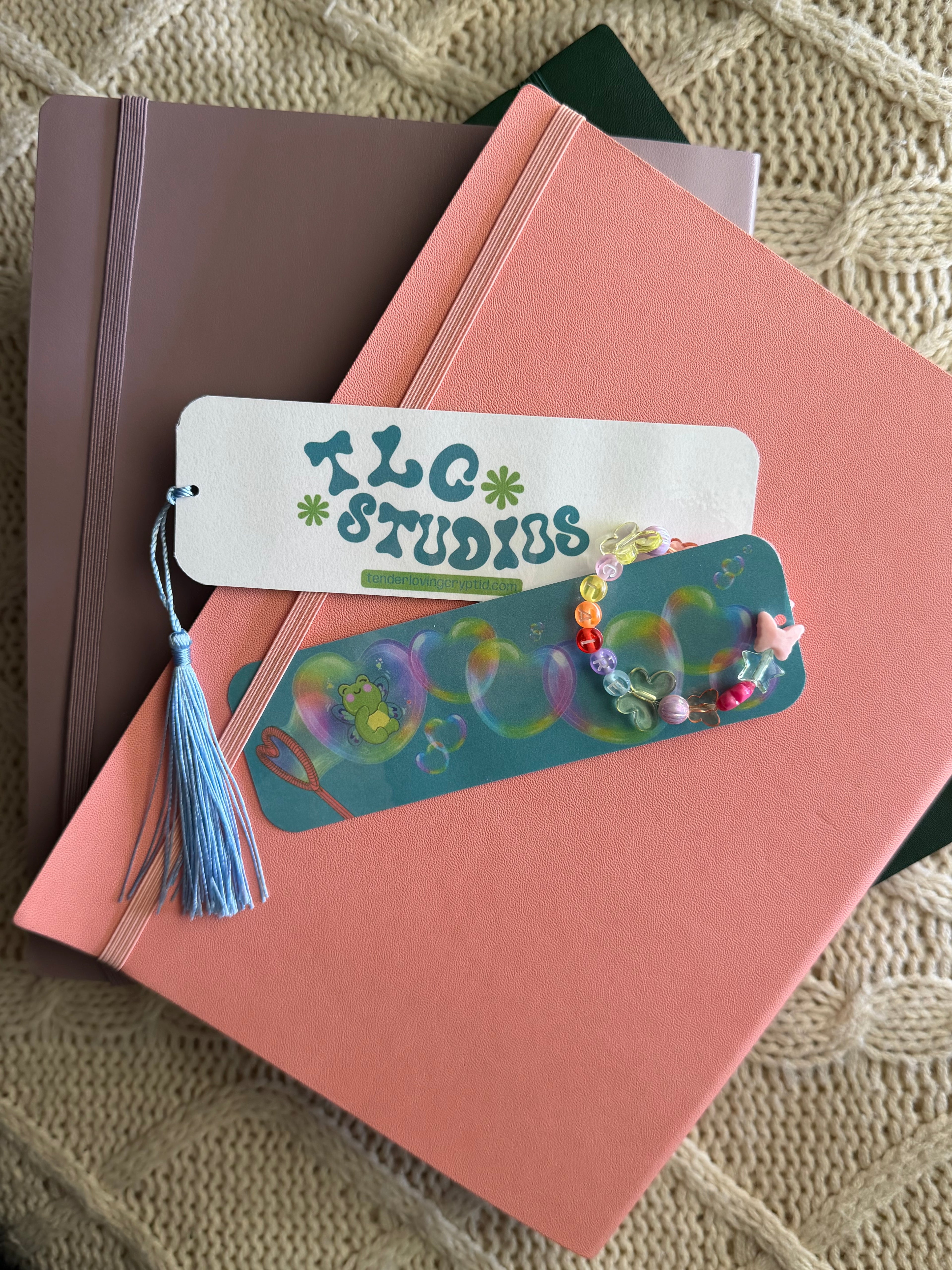 Two bookmarks, with one featuring the art print that covers the front while the other shows the back that reads 'TLC STUDIOS' in teal, placed on top of stacked notebooks on a beige knit blanket.