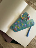 A pair of decorative bookmarks with a blue tassel and a matching beaded bracelet, both featuring a colorful bubble pattern, resting on an open lined notebook.