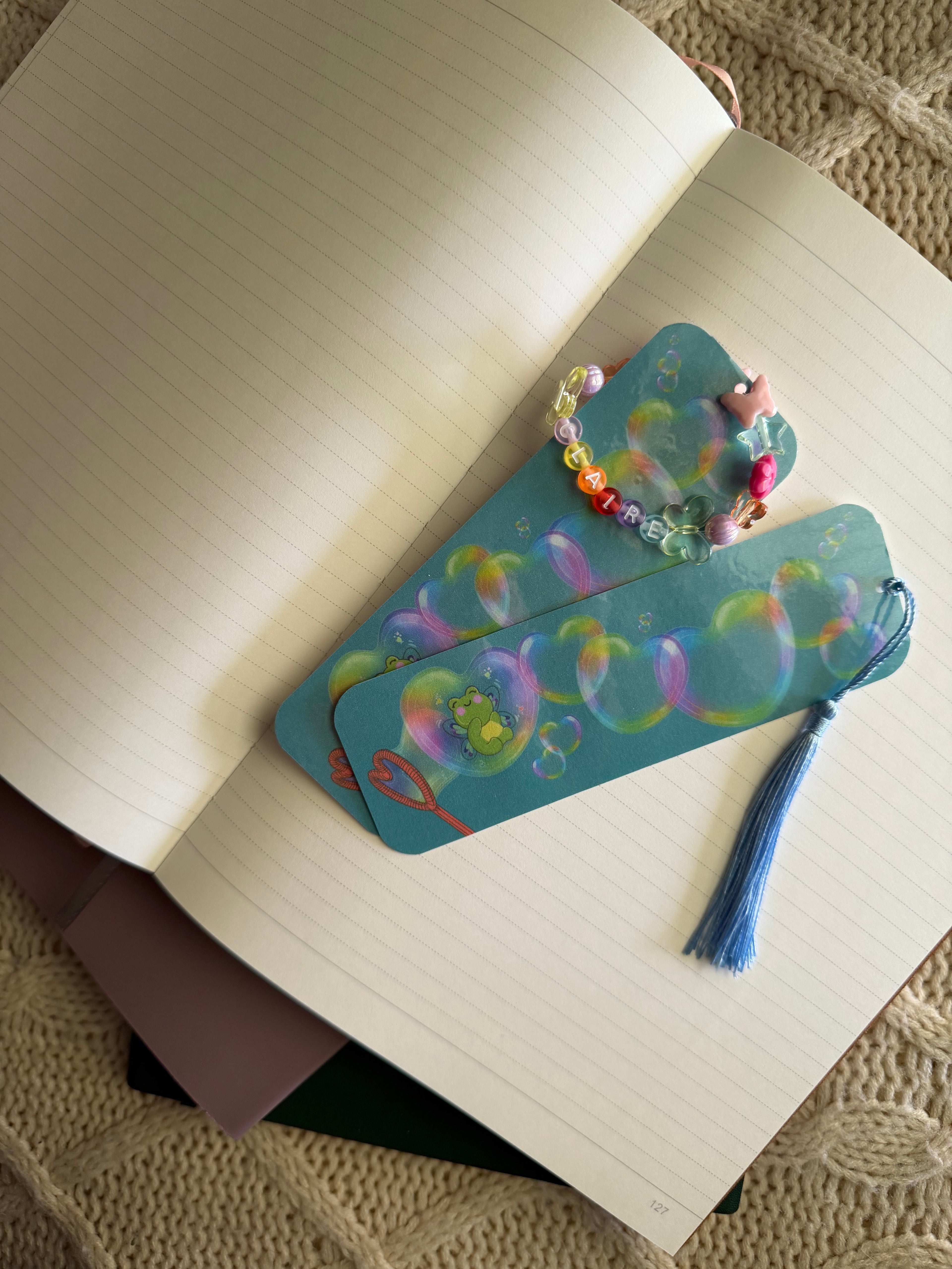 A pair of decorative bookmarks with a blue tassel and a matching beaded bracelet, both featuring a colorful bubble pattern, resting on an open lined notebook.